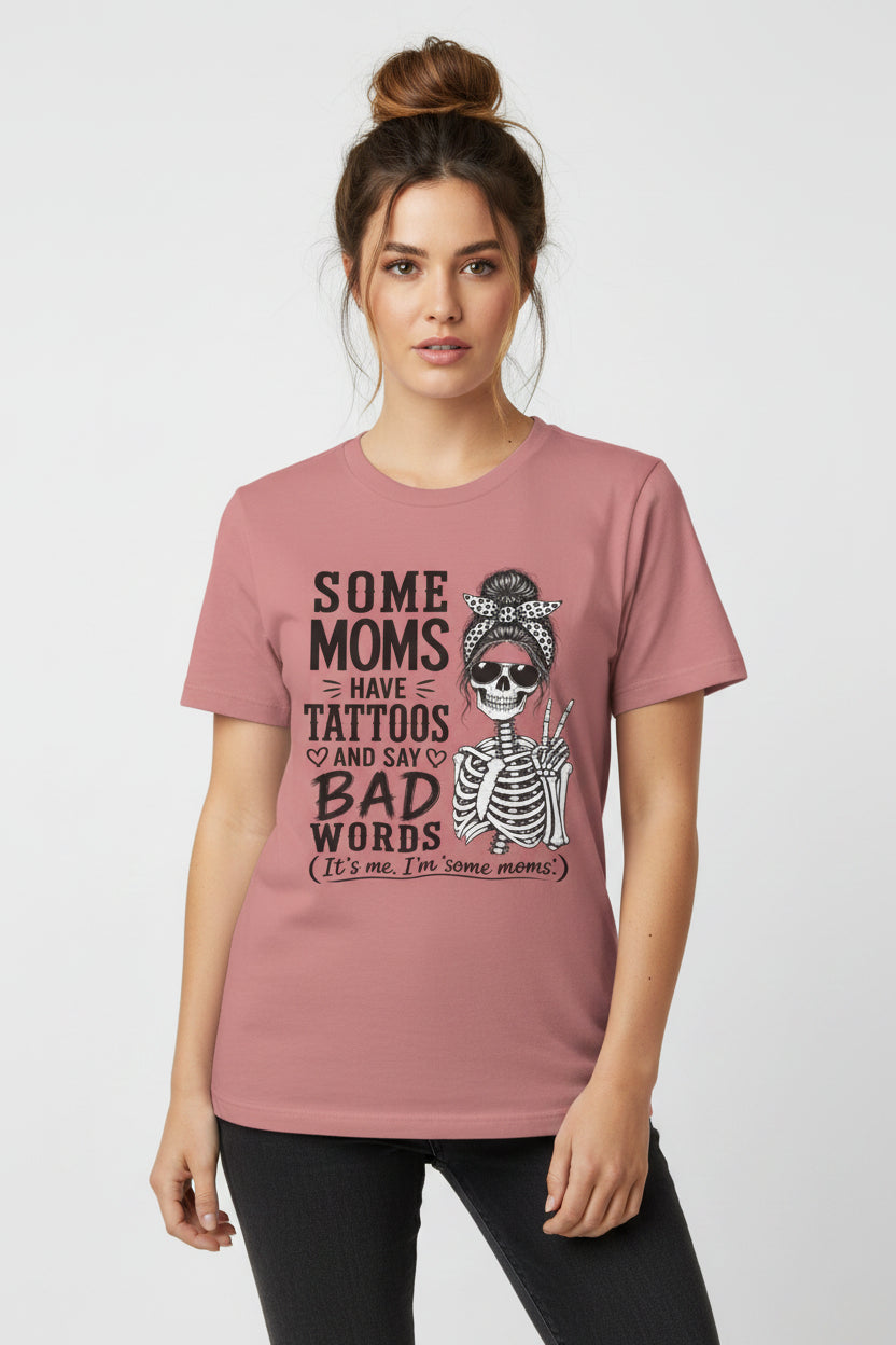 A dusty rose short-sleeve t-shirt with a graphic print featuring a skeleton and the text 'SOME MOMS HAVE TATTOOS AND SAY BAD WORDS (It's me, I'm some moms.)' on a solid white background.