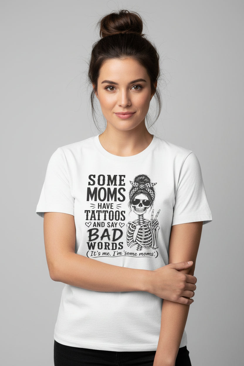 A white short-sleeve crewneck t-shirt featuring a black graphic print of a skeleton woman with a bandana and sunglasses, next to the text 'SOME MOMS HAVE TATTOOS AND SAY BAD WORDS (It's me, 'some moms.)'.